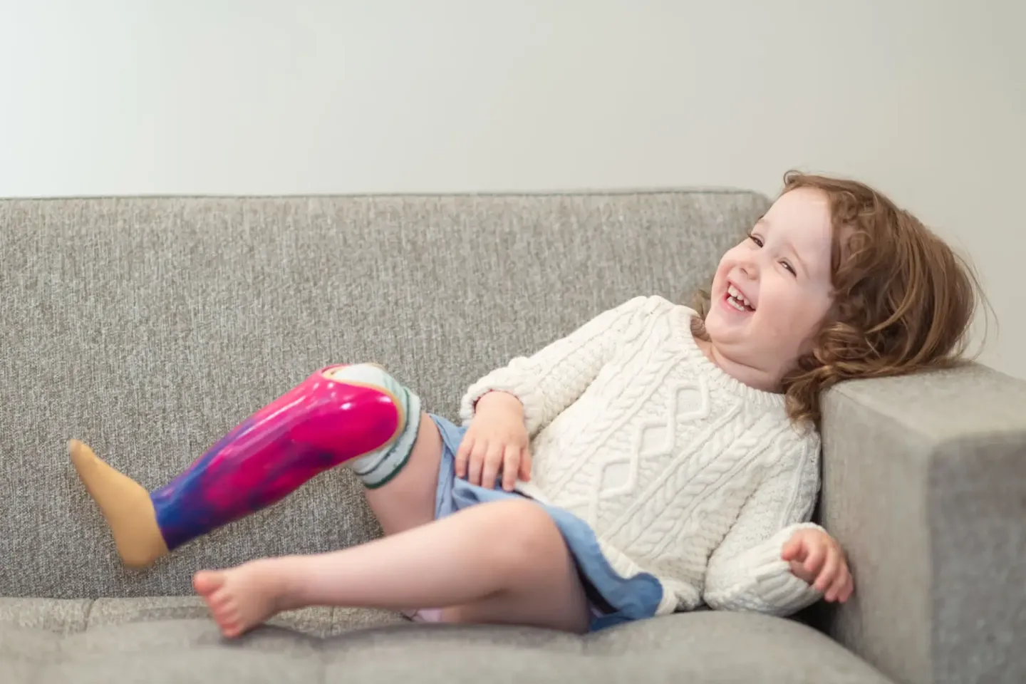 A girl with a pink prosthetic leg laughs on a couch.